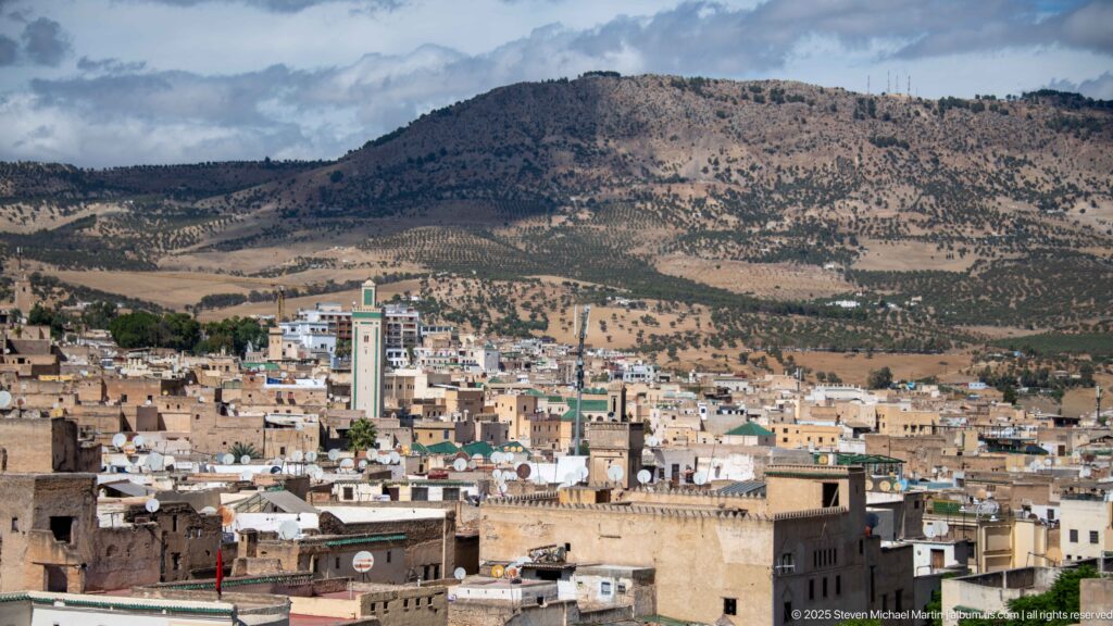 Panoramic view over Fes rooftops and minarets toward a broad dry mountain ridge.