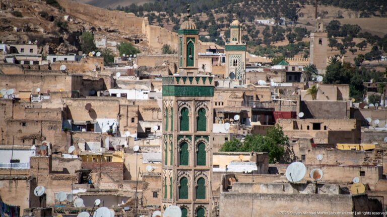 Green-decorated square minaret rising above dense Fes rooftops with satellite dishes and other towers behind.