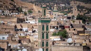 Green-decorated square minaret rising above dense Fes rooftops with satellite dishes and other towers behind.