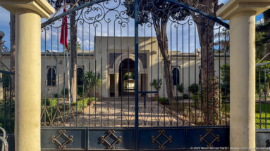 Ornate wrought-iron gate framing a formal courtyard with palms and a pale building beyond.