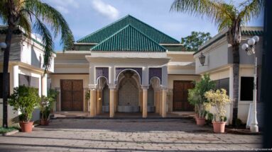 Courtyard pavilion with green-tiled roof, central arched fountain niche, paired columns, wooden doors, and potted plants.