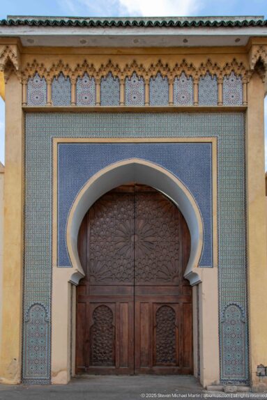Large carved wooden double door beneath a horseshoe arch framed by blue and turquoise tilework.