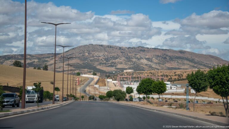 Wide boulevard with cars and streetlights descending toward rocky hills and developing outskirts of Fes.