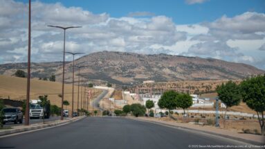 Wide boulevard with cars and streetlights descending toward rocky hills and developing outskirts of Fes.