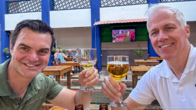 Two smiling men raising beer glasses at an outdoor terrace with wooden tables and a television behind them.