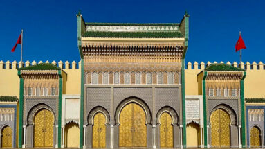 Front view of Royal Palace gates with brass doors, green roof tiles, flanking portals, crenellated walls, and Moroccan flags.