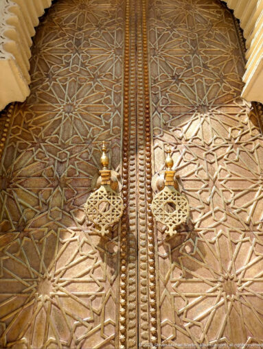 Close-up of a brass palace door with two ornate ring handles and dense interlaced geometric star patterns.