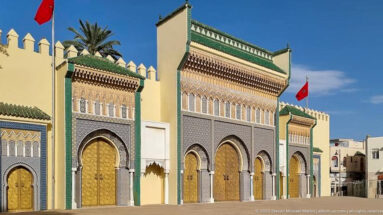 Oblique view of Royal Palace gates with brass doors, patterned façades, green-tiled roofs, crenellations, and Moroccan flag.