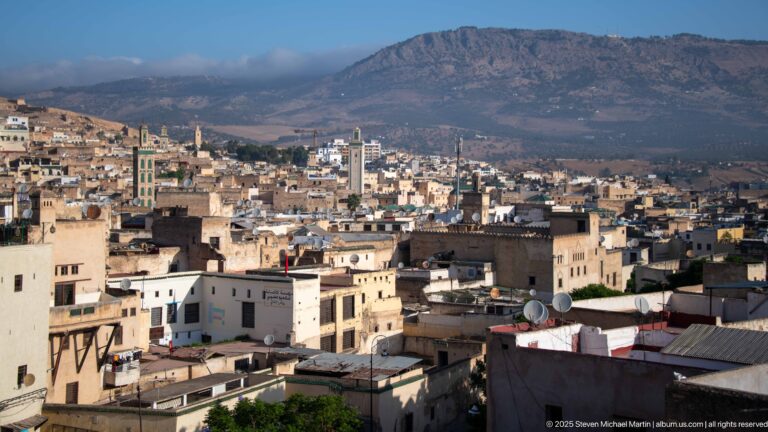 Panoramic view over Fes rooftops and minarets toward a broad mountain ridge under a clear sky.