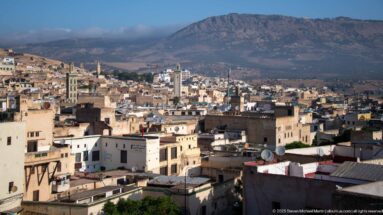 Panoramic view over Fes rooftops and minarets toward a broad mountain ridge under a clear sky.