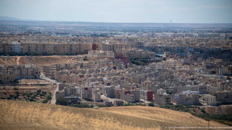 Golden field overlooking dense low-rise neighborhoods on the outskirts of Fes stretching toward the horizon.