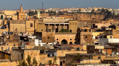 Dense Fes medina rooftops with earthen walls, laundry, satellite dishes, minaret, and a large arched building façade.