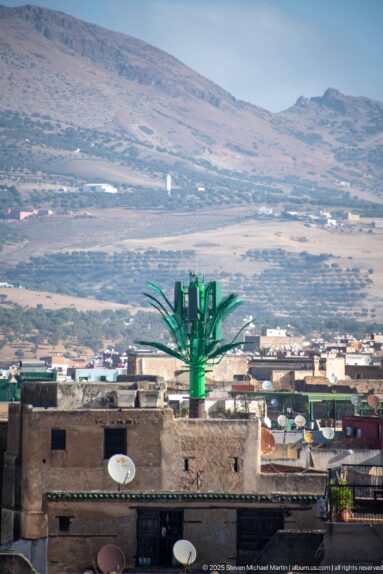 Elevated Fes rooftops with satellite dishes, a bright green metal structure, and a broad dry hillside in the distance.