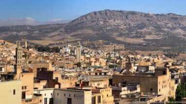 Elevated view of Fes with dense ochre rooftops, several minarets, and a dry rocky mountain ridge behind.