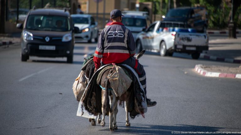 Man riding a loaded donkey along a city road in Fes with cars traveling around him.