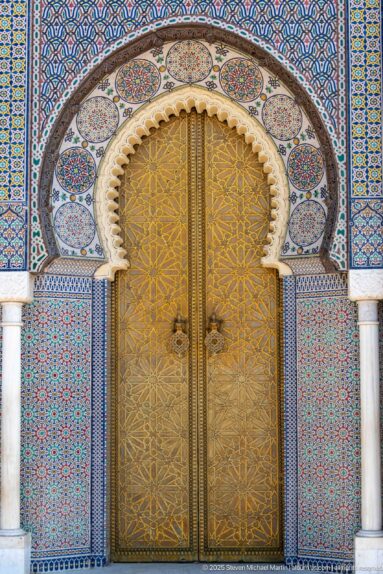 Close view of a brass palace door beneath a scalloped arch framed by multicolored zellij and white columns.