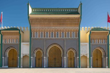 Symmetrical Royal Palace façade with brass doors, ornate arches, green-tiled roofs, crenellated walls, and Moroccan flags.