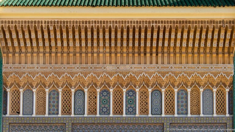 Long palace façade detail with narrow arches, multicolored zellij, carved stucco, projecting wooden eaves, and green roof tiles.