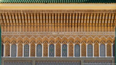 Long palace façade detail with narrow arches, multicolored zellij, carved stucco, projecting wooden eaves, and green roof tiles.