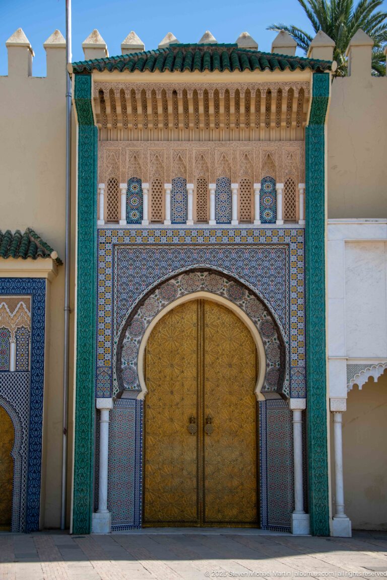 Ornate brass double door beneath a horseshoe arch with zellij, white columns, turquoise panels, and green roof tiles.