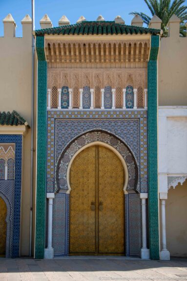 Ornate brass double door beneath a horseshoe arch with zellij, white columns, turquoise panels, and green roof tiles.