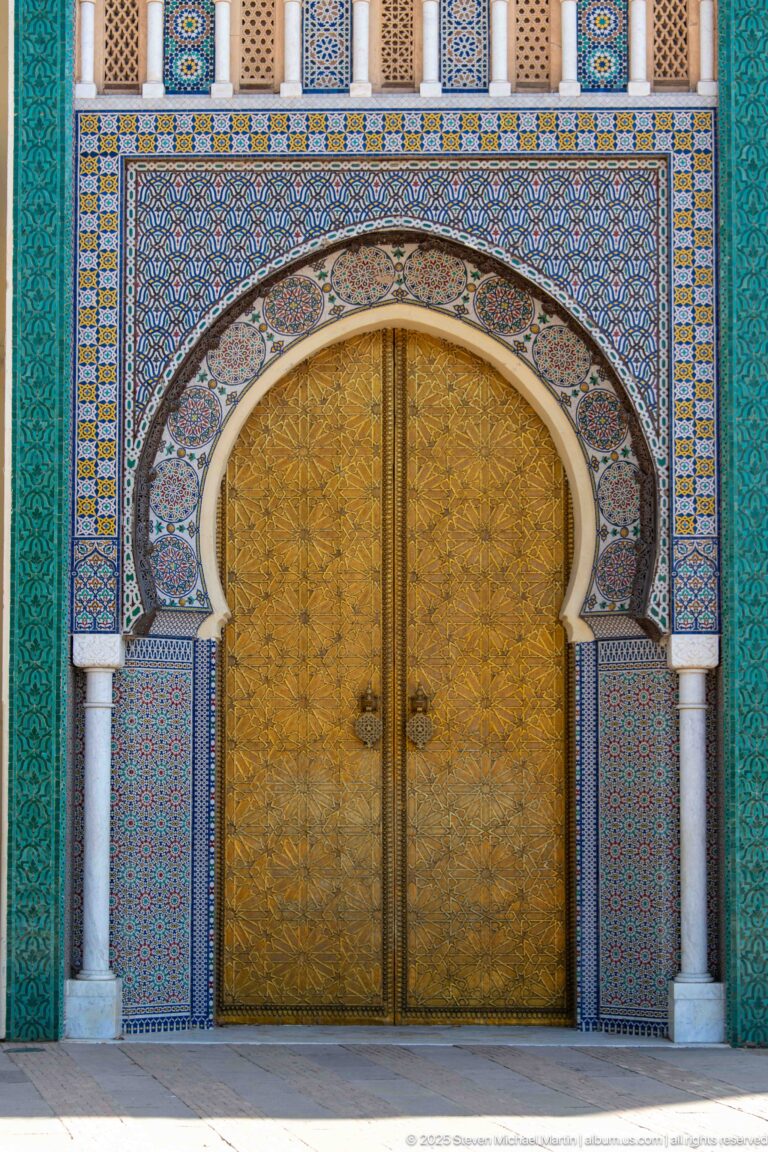 Front view of a monumental brass double door framed by horseshoe arch, zellij, columns, and turquoise panels at the Royal Palace of Fes.