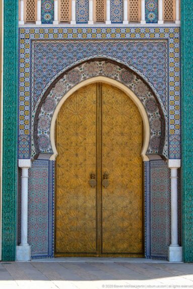 Front view of a monumental brass double door framed by horseshoe arch, zellij, columns, and turquoise panels at the Royal Palace of Fes.