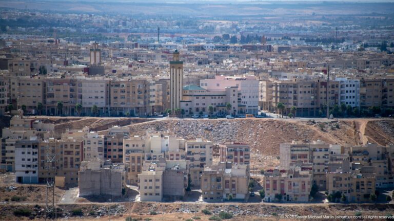 Modern apartment blocks and construction zones on the outskirts of Fes, with a tall tower and dense development beyond.