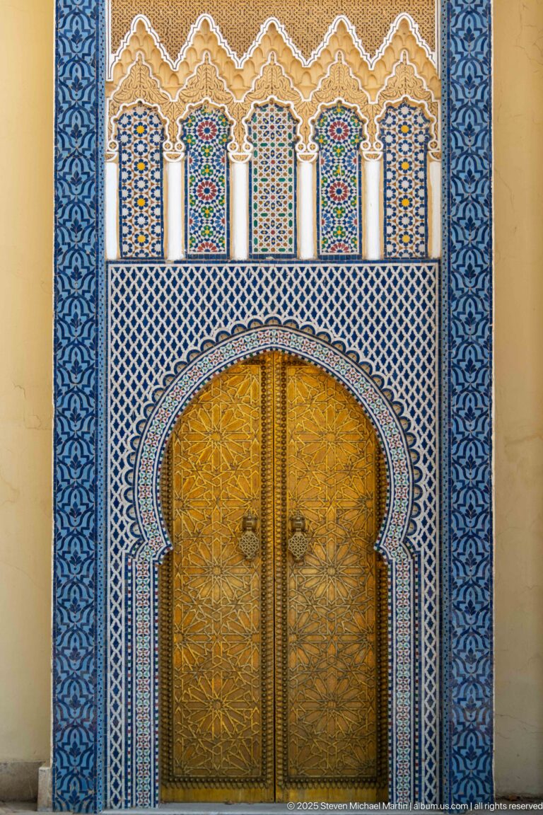 Large brass double door beneath a horseshoe arch surrounded by blue-white zellij and carved decoration at the Royal Palace of Fes.
