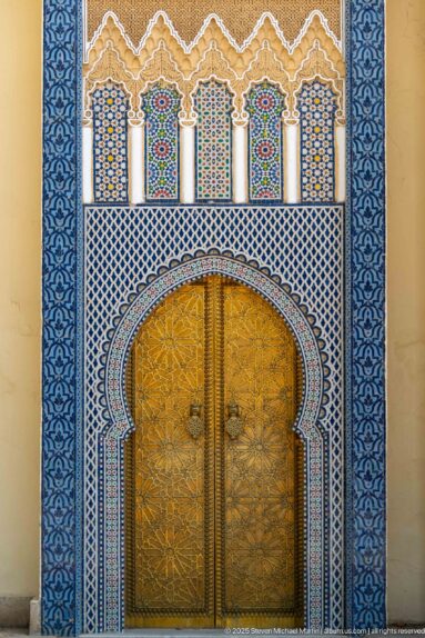 Large brass double door beneath a horseshoe arch surrounded by blue-white zellij and carved decoration at the Royal Palace of Fes.
