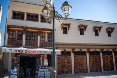 White building in Fes with wooden lattice windows, carved timber details, tiled eaves, shopfront, closed wooden doors, and ornate lamps.