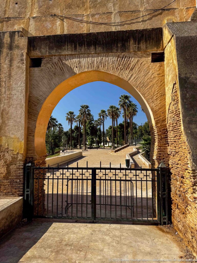 Large masonry arch and black metal gate opening onto a sunlit palm-lined garden path in Fes.