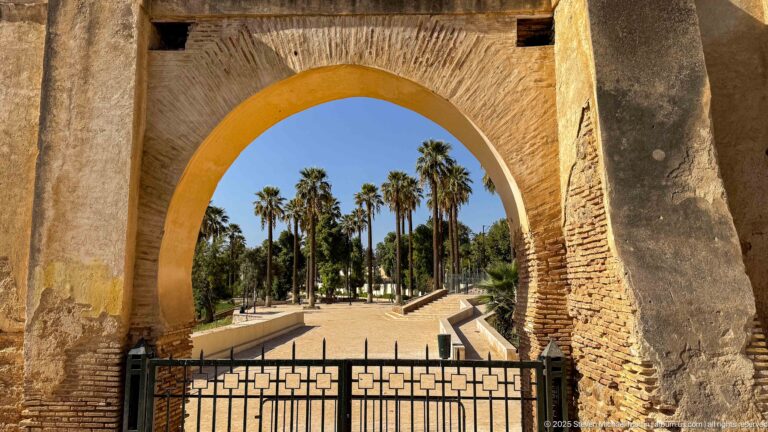 Large brick-and-plaster arch framing a gated palm-lined garden path in the Royal Palace precinct of Fes.