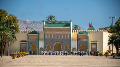 Symmetrical ceremonial entrance to the Royal Palace of Fes with green roofs, arched gateways, crowds, palms, and mountains.