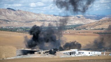 Thick black smoke rising from low buildings in farmland outside Fes, with dry fields and pale mountains beyond.