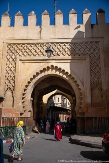 Scalloped horseshoe gate with carved geometric panels and crenellations, opening onto a busy street in Fes.