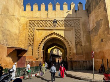 Sunlit ochre gate with scalloped arch, geometric carved panels, crenellations, and pedestrians entering the medina.