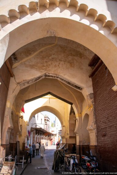 Series of broad arches forming a shaded passage into a busy Fes street with pedestrians and stacked goods.