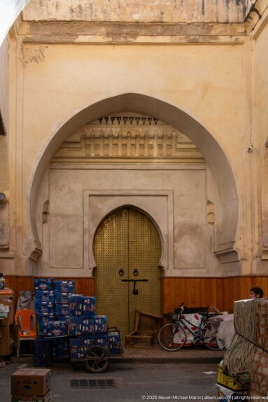 Large arched street fountain or niche with metal grille, stacked goods, handcart, and bicycles along the wall.