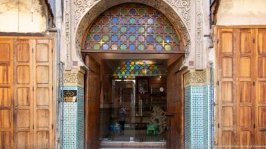 Decorated shop entrance with carved arch, turquoise tilework, geometric grille, wooden shutters, and merchandise inside.
