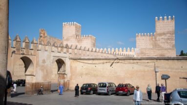 Historic crenellated walls and square towers in Fes above parked cars and pedestrians along the street.