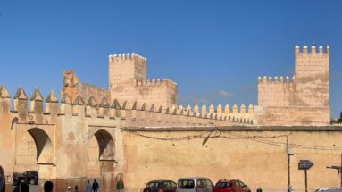 Layered crenellated walls and square defensive towers in Fes with parked cars and utility lines below.