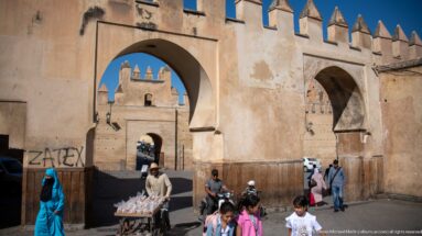 Busy street beneath large arches in the walls of Fes, with pedestrians, vendor cart, cars, crenellations, and a square tower.
