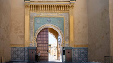 Pointed ceremonial gateway with blue-green geometric tilework, zellij panels, and a partly open wooden door.