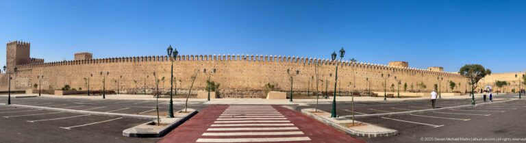 Long ochre crenellated wall in Fes beside a broad roadway and parking area under a clear blue sky.