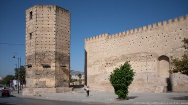 Massive rectangular masonry tower beside a high fortified wall in Fes, with a person standing at the base for scale.
