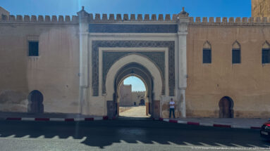 Large pointed gateway in an ochre crenellated wall, framed with geometric ornament and flanked by smaller arched openings.