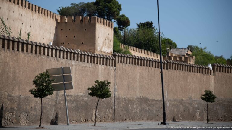 Pinkish crenellated wall and square tower in Fes, with small street trees, road signs, and taller trees behind.