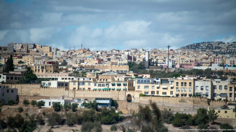 Wide view of dense hillside neighborhoods in Fes beyond a lower masonry wall and pale urban buildings.