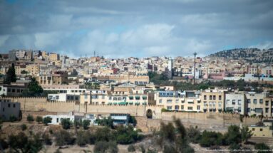 Wide view of dense hillside neighborhoods in Fes beyond a lower masonry wall and pale urban buildings.